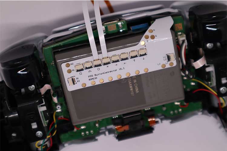 Nahaufnahme der internen Komponenten eines PS5 Remapper V2 BDM030/040/050, mit einer Platine, an der Flachbandkabel befestigt sind, und einer Batterie mit der Aufschrift "Made in China" darunter.