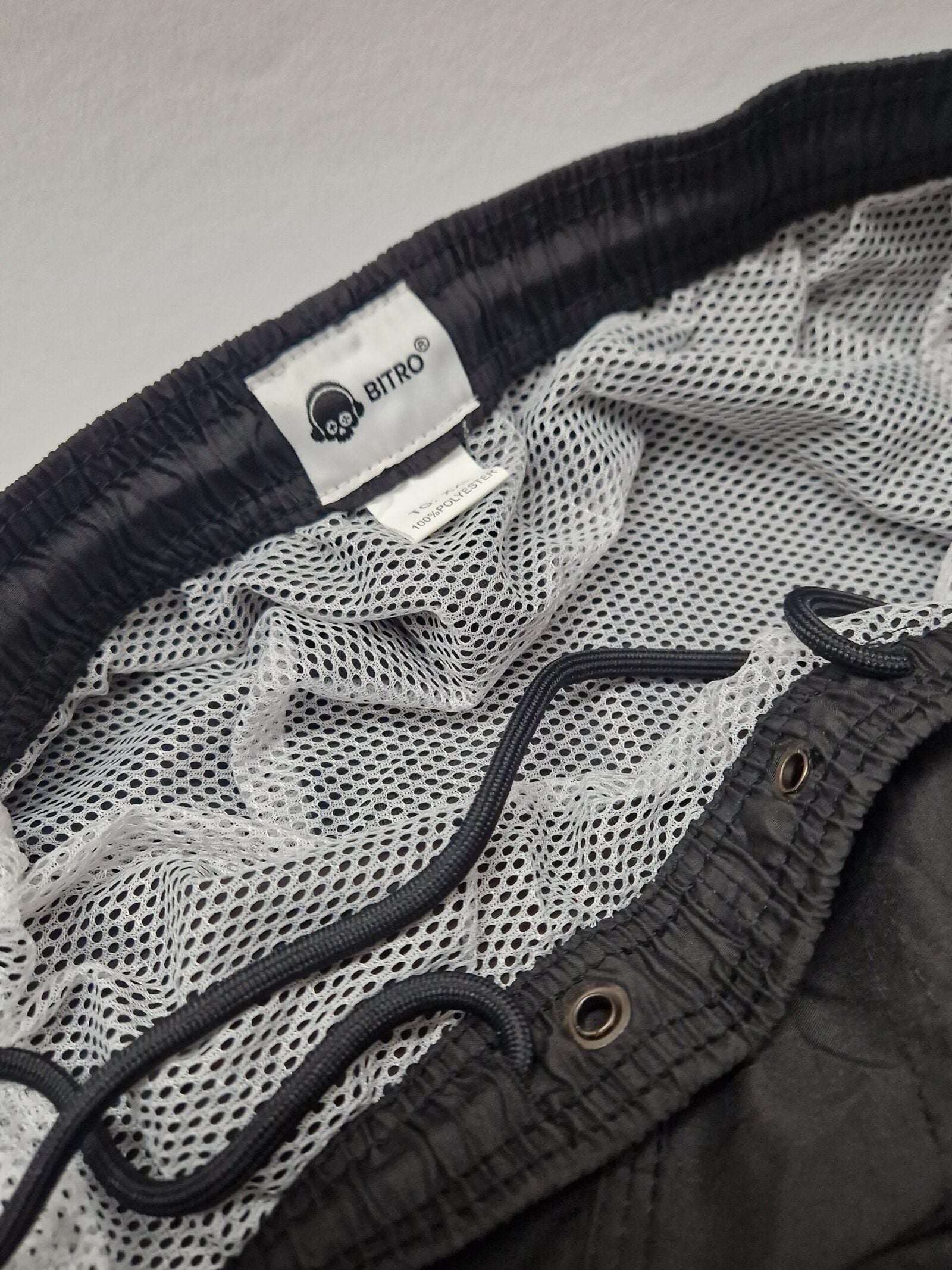 Nahaufnahme des Innenbundes einer schwarzen Shorts mit weißem Mesh-Futter und einem schwarzen Kordelzug. In den Bund ist ein Etikett mit dem Markennamen BITRO und einem Logo eingenäht.